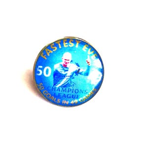 May include: A round, blue pin with gold lettering and a picture of a soccer player. The pin reads "FASTEST EVER" and "50 CHAMPIONS LEAGUE GOALS IN 49 GAMES". The soccer player is wearing a blue jersey.