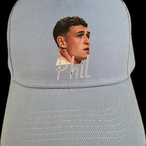 May include: Light blue baseball cap featuring a printed image of a person's face and the name "Phil" in white script. The cap has a curved brim and an adjustable strap at the back, suitable for casual wear.