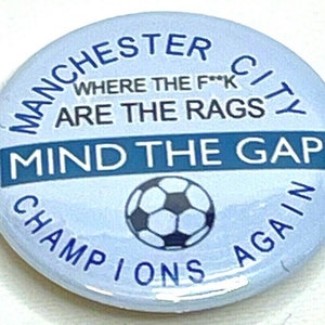 May include: A light blue button with the text "MANCHESTER CITY" and "WHERE THE F**K ARE THE RAGS". It also says "MIND THE GAP" and "CHAMPIONS AGAIN". A soccer ball graphic is in the center.