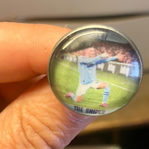 May include: A round, silver-colored button with a clear, domed surface. The button features a photograph of a soccer player in a light blue uniform on a green field. The text "THE SNIPER" is printed at the bottom.