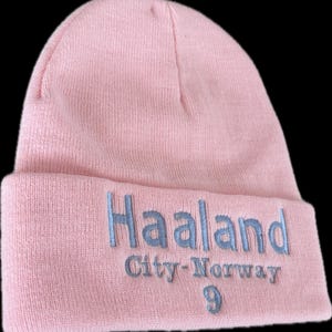 以下が含まれることがあります： 薄いピンク色のニット帽。ライトブルーで「Haaland」、「City-Norway」、数字の「9」が刺繍されています。柔らかいニット素材でできており、折り返しのついたつばがあります。