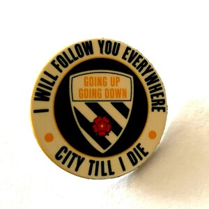 以下が含まれることがあります： クリーム色の縁取りに黒文字で「I WILL FOLLOW YOU EVERYWHERE」および「CITY TILL I DIE」と書かれた円形のエナメルピン。中央には黒い斜めストライプ、赤いバラ、そして「GOING UP GOING DOWN」という文字が描かれた盾の紋章が配置されています。