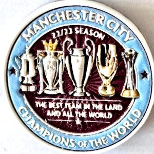 以下が含まれることがあります： マンチェスターシティの丸い記念コイン。コインには「Manchester City 22/23 Season Champions of the World」の文字。金と銀のトロフィーのイラストが、明るい青色の縁取りのあるバーガンディ色の背景に描かれています。
