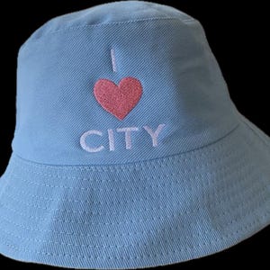 以下が含まれることがあります： 「I ❤️ CITY」の刺繍が入った水色のバケットハット。ハートはピンク色、文字は白色です。柔らかい生地で作られており、クラシックなバケットハットの形をしています。