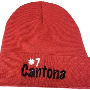 Puede incluir: Un gorro de punto rojo brillante con un borde doblado. El gorro presenta el texto bordado "#7 Cantona" en hilo negro y blanco. El gorro está hecho de un material de punto suave.