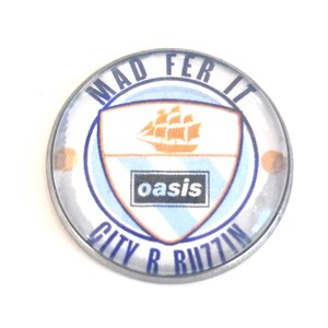 Puede incluir: Un botón redondo con borde plateado, con el texto "MAD FER IT" y "CITY R BUZZIN" en azul. El centro presenta un escudo con un barco y la palabra "oasis". El fondo es blanco.