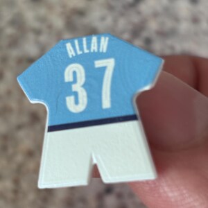 Puede incluir: Un pequeño pin o ficha plano con forma de camiseta deportiva. La parte superior es de color azul claro con el nombre ALLAN y el número 37 impresos en blanco. Una fina línea horizontal azul oscuro separa la parte superior azul claro de la base blanca.