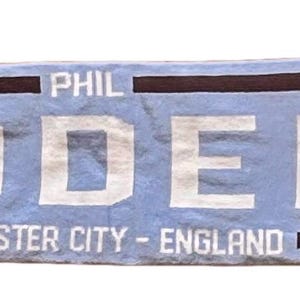 May include: A light blue and white scarf with the name "PHIL FODEN" in large white letters. The scarf has a portrait of a person on each end and the text "MANCHESTER CITY - ENGLAND". The scarf has white fringe.