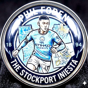 May include: A round, silver-toned metal pin featuring a stylized illustration of a soccer player in a light blue jersey. The pin has the text "PHIL FODEN" and "THE STOCKPORT INIESTA" around the edge. The player is depicted in action, with a map-like background.