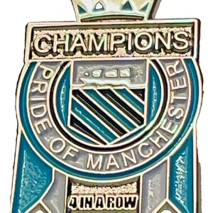 May include: A silver and teal enamel pin with a crown at the top. The pin reads "CHAMPIONS" and "PRIDE OF MANCHESTER" around a shield design. The bottom of the pin says "4 IN A ROW".