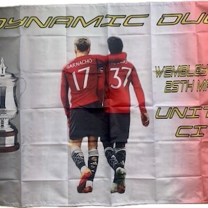 Puede incluir: Bandera con un diseño degradado negro, blanco y rojo. Presenta el texto "DYNAMIC DUO" y "WEMBLEY STADIUM 25TH MAY 2024". La bandera también muestra el marcador "UNITED 2 CITY 1" e imágenes de dos futbolistas.