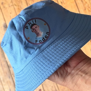 May include: Light blue bucket hat featuring a circular graphic with the name "Phil Foden" and a portrait of a young man. The hat is made of a soft fabric and has a classic bucket hat design.