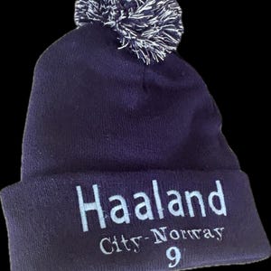 Puede incluir: Un gorro de punto azul marino con un pompón blanco y azul marino. El borde doblado muestra el nombre "Haaland", "City-Norway" y el número "9" en letras blancas.