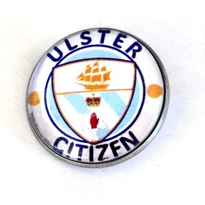 Puede incluir: Un botón redondo con borde plateado, con las palabras "ULSTER CITIZEN" en azul. El botón presenta un diseño de escudo con un barco, una corona y una mano roja sobre un fondo blanco y azul.