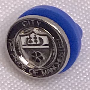 Puede incluir: Pin de solapa azul y plateado con las palabras "CITY" y "THE PRIDE OF MANCHESTER" alrededor de un escudo. El escudo tiene forma de escudo con un árbol y una rueda.