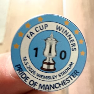 May include: A round, light blue badge with the text "FA CUP WINNERS" and "PRIDE OF MANCHESTER". The badge features a silver trophy and the date "16.5.2026". Gold stars encircle the badge.