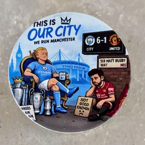 Puede incluir: Una pegatina circular con una caricatura de un futbolista con camiseta azul claro sentado en un trono con trofeos, junto a un jugador con camiseta roja sentado en el suelo. El texto incluye THIS IS OUR CITY, WE RUN MANCHESTER y 6-1.