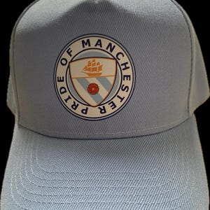 Op de afbeelding: Lichtblauwe baseballcap met het Manchester City-embleem. Het embleem is een cirkelvormig ontwerp met een schip, een roos en de woorden "Pride of Manchester". De cap heeft een gebogen klep en een gestructureerde kroon.