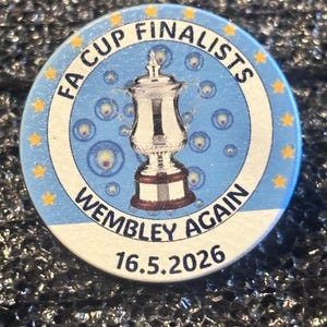 以下が含まれることがあります： 「FA CUP FINALISTS WEMBLEY AGAIN 16.5.2026」の文字が入った、丸い青と白のピンバッジ。中央には銀色のトロフィーがあり、星と泡で囲まれています。