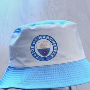 May include: A light blue and white bucket hat featuring a circular emblem on the front. The emblem contains the text PRIDE OF MANCHESTER surrounding a shield graphic that displays a ship above a red flower on a blue and white background.