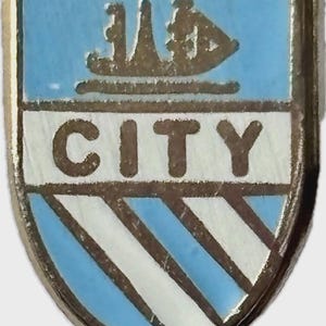 Puede incluir: Pin de esmalte vintage con diseño de escudo en azul claro y blanco. La palabra "CITY" se muestra en blanco, con un fondo azul claro arriba y rayas diagonales abajo. Un emblema dorado en la parte superior.
