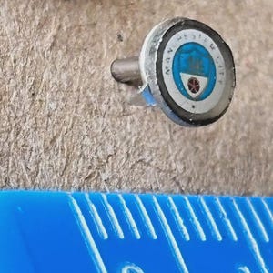 Puede incluir: Un pequeño pin redondo con el logotipo del Manchester City. El pin presenta un diseño azul y blanco con las palabras "MANCHESTER CITY" alrededor del borde. El pin está unido a un poste plateado.
