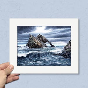 May include: A framed watercolor painting depicts a dramatic coastal scene. A large rock formation with an arch stands in the turbulent sea under a stormy sky. The waves crash against the rocks, creating a dynamic and moody seascape.