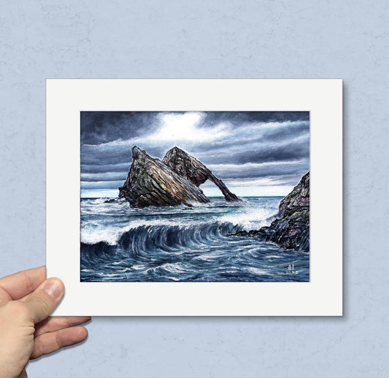 May include: A framed watercolor painting depicts a dramatic coastal scene. A large rock formation with an arch stands in the turbulent sea under a stormy sky. The waves crash against the rocks, creating a dynamic and moody seascape.