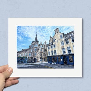 May include: A framed print depicting a street scene with historic buildings under a blue sky. The buildings are made of stone and have blue accents. The print is held by a hand.