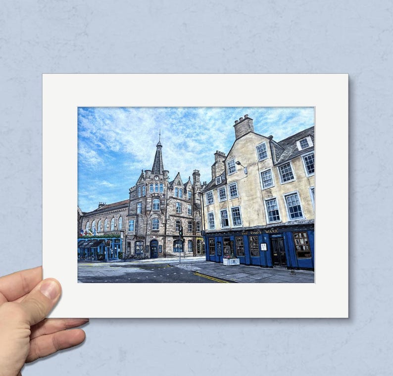 May include: A framed print depicting a street scene with historic buildings under a blue sky. The buildings are made of stone and have blue accents. The print is held by a hand.
