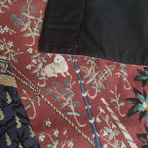 May include: Close-up of a tapestry fabric with a red background and a black border. The tapestry features a detailed design of flowers, leaves, and birds in shades of green, blue, and white.