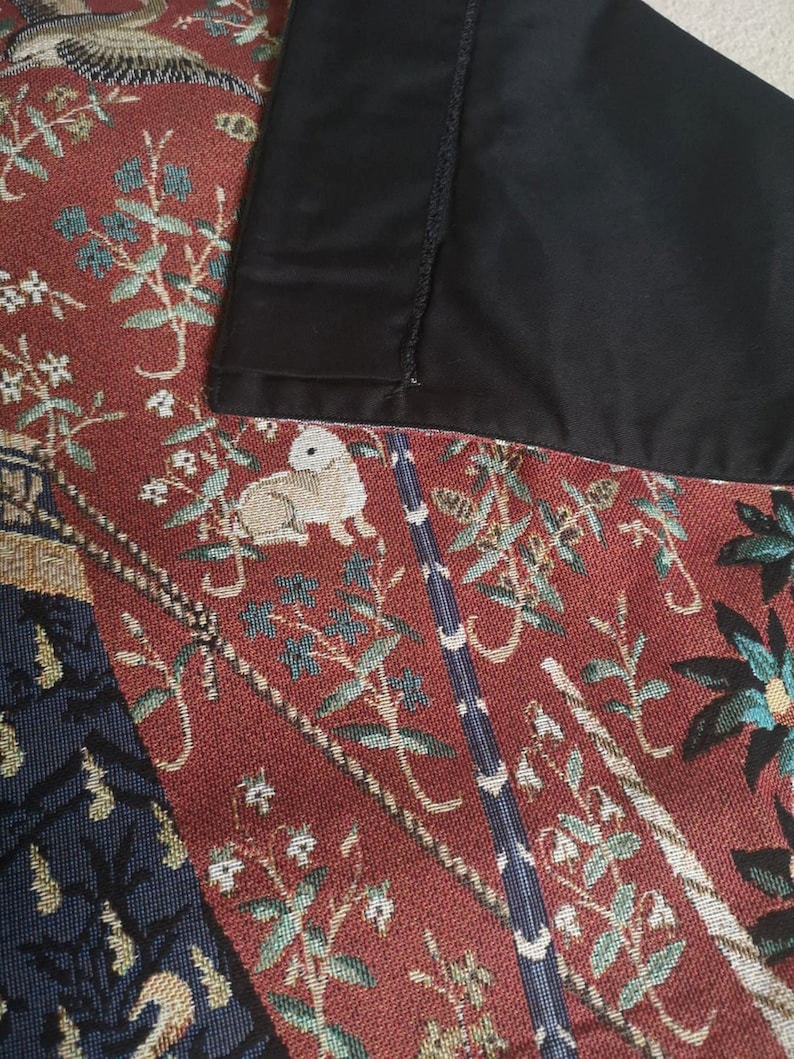 May include: Close-up of a tapestry fabric with a red background and a black border. The tapestry features a detailed design of flowers, leaves, and birds in shades of green, blue, and white.