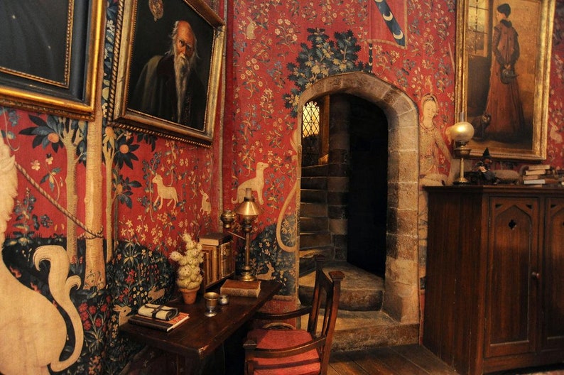 May include: A red and gold tapestry with a unicorn design hangs on the wall of a room with a wooden desk, chair, and a doorway leading to a staircase.