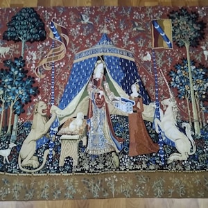May include: A tapestry depicting a unicorn kneeling before a lady in a blue and gold tent. The unicorn is white with a gold horn and a blue and gold bridle. The lady is wearing a red and gold dress and a blue and gold crown. The tapestry is surrounded by a border of flowers and leaves.