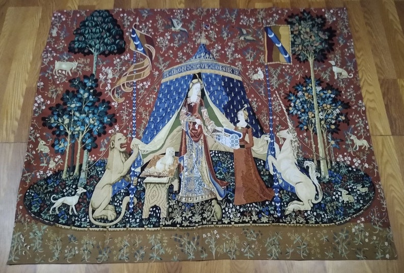May include: A tapestry depicting a unicorn kneeling before a lady in a blue and gold tent. The unicorn is white with a gold horn and a blue and gold bridle. The lady is wearing a red and gold dress and a blue and gold crown. The tapestry is surrounded by a border of flowers and leaves.