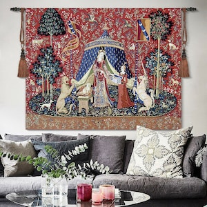 May include: A tapestry featuring a red background with a scene of a woman in a blue tent with a unicorn and a lion. The tapestry has a medieval theme and is framed with tassels.