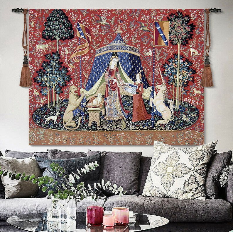 May include: A tapestry featuring a red background with a scene of a woman in a blue tent with a unicorn and a lion. The tapestry has a medieval theme and is framed with tassels.
