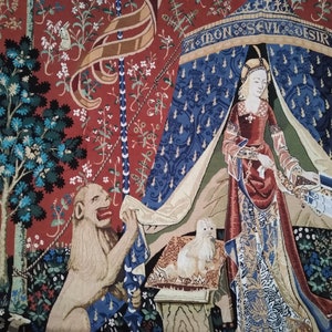 May include: A tapestry depicting a scene from the Lady and the Unicorn series. A woman in a red and blue gown stands under a canopy with the text "A mon seul desir". A lion with a red mane sits on the ground to the left of the woman. The tapestry is woven with intricate details and vibrant colors.