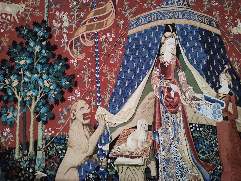 May include: A tapestry depicting a scene from the Lady and the Unicorn series. A woman in a red and blue gown stands under a canopy with the text "A mon seul desir". A lion with a red mane sits on the ground to the left of the woman. The tapestry is woven with intricate details and vibrant colors.