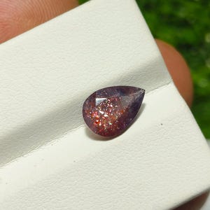 May include: A teardrop-shaped gemstone with a purple and red iridescent sheen. The gemstone is set on a white background.