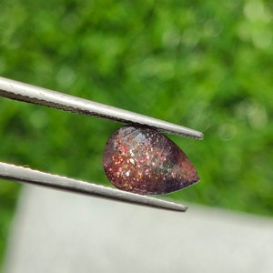 May include: A pear-shaped gemstone with a reddish-brown color and a glittery, iridescent finish. The gemstone is held by a pair of tweezers.