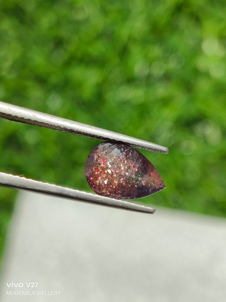 May include: A pear-shaped gemstone with a reddish-brown color and a glittery, iridescent finish. The gemstone is held by a pair of tweezers.
