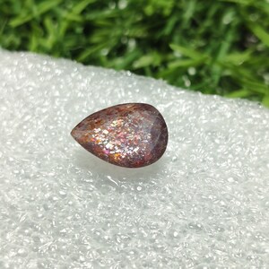 May include: A teardrop-shaped gemstone with a reddish-brown base and iridescent sparkle. The gemstone is set against a white background.