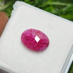 Puede incluir: Una gema de rubí ovalada y facetada. La gema es de un color rojo rosado vibrante, con una faceta reflectante prominente en el centro. Descansa sobre una superficie blanca dentro de una caja de plástico transparente, con un fondo verde borroso.