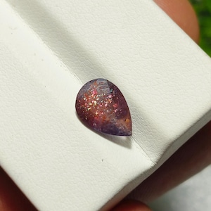 May include: A teardrop-shaped gemstone with a purple and pink hue. The gemstone has a sparkly, iridescent finish.
