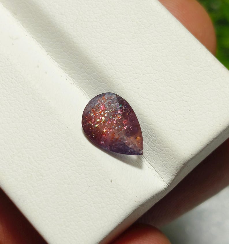 May include: A teardrop-shaped gemstone with a purple and pink hue. The gemstone has a sparkly, iridescent finish.