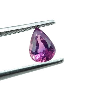 Puede incluir: Una gema facetada en forma de pera, en tonos rosa y púrpura, es sostenida por unas pinzas plateadas. La gema tiene un alto grado de claridad y brillo, con una superficie pulida que refleja la luz. El fondo es blanco.