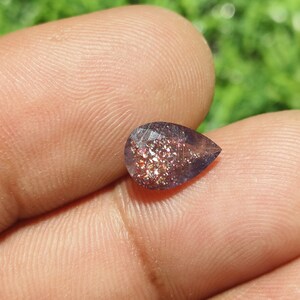May include: A pear-shaped gemstone with a purple hue and a glittery, iridescent finish. The stone is set against a blurred background of green foliage.