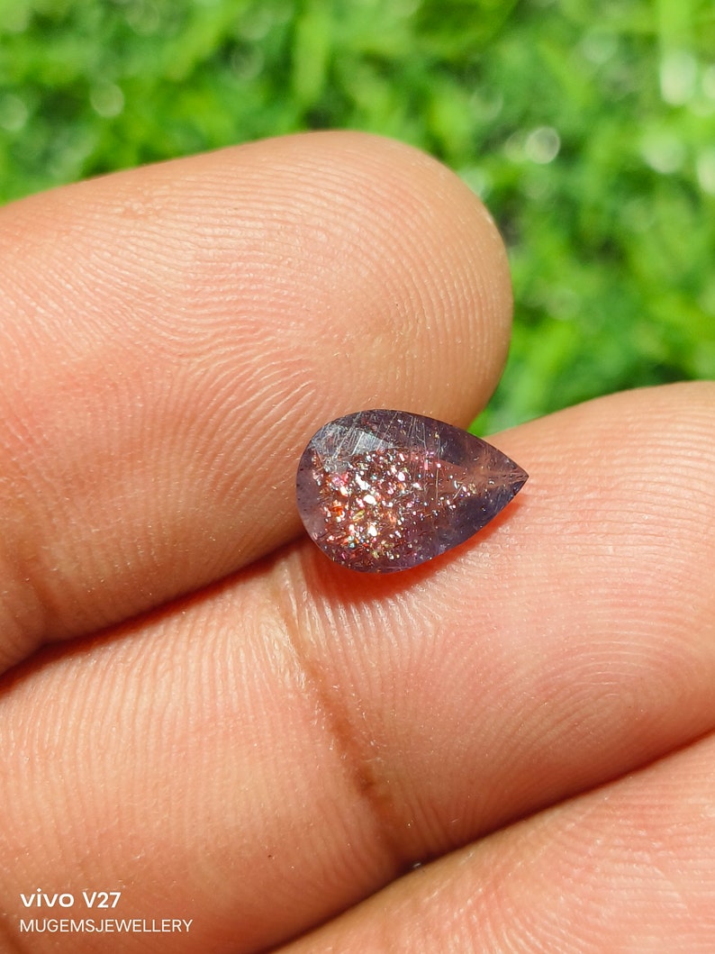 May include: A pear-shaped gemstone with a purple hue and a glittery, iridescent finish. The stone is set against a blurred background of green foliage.