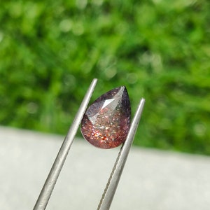 May include: A pear-shaped gemstone with a reddish-brown hue and iridescent sparkle. The gemstone is held by tweezers against a blurred green background.